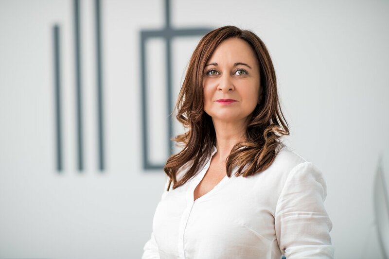 Portrait von Iris Radakovits in weißer Bluse vor einer weißen Wand mit Firmenlogo